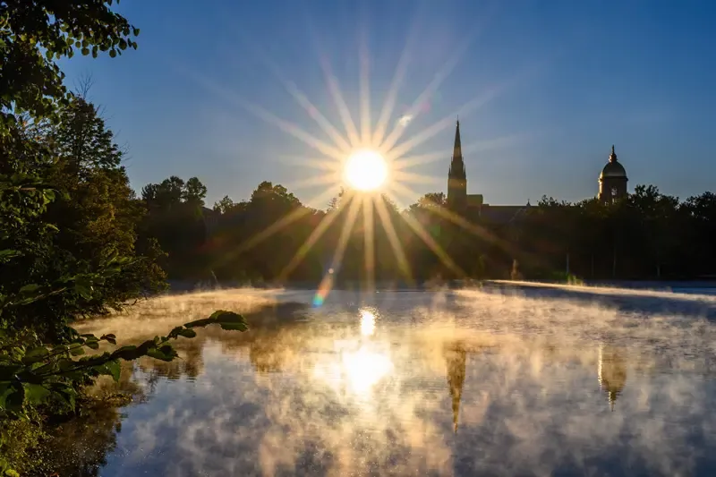 The sun rises over St. Mary's Lake on the University of Notre Dame campus. The Golden Dome and Basilica of the Sacred Heart are silhouetted against the brilliant sunburst. Wisps of fog drift across the lake, reflecting the light and the iconic landmarks. Green trees frame the right side of the image.